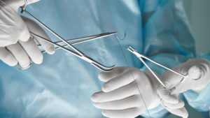 Two doctors, dressed in disposable surgery scrubs and gloves, prepare a suture. They use a needle holder, toothed forceps, fine suturing scissors, and suturing material.