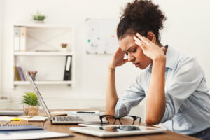 Woman sitting at her desk in front of her computer with her eyes closed touching her temples with both hands in pain.