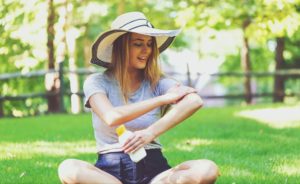 A woman in a hat and summer clothes sits on the grass, applying sunscreen to her arm with one hand while holding a sunscreen bottle in the other hand.