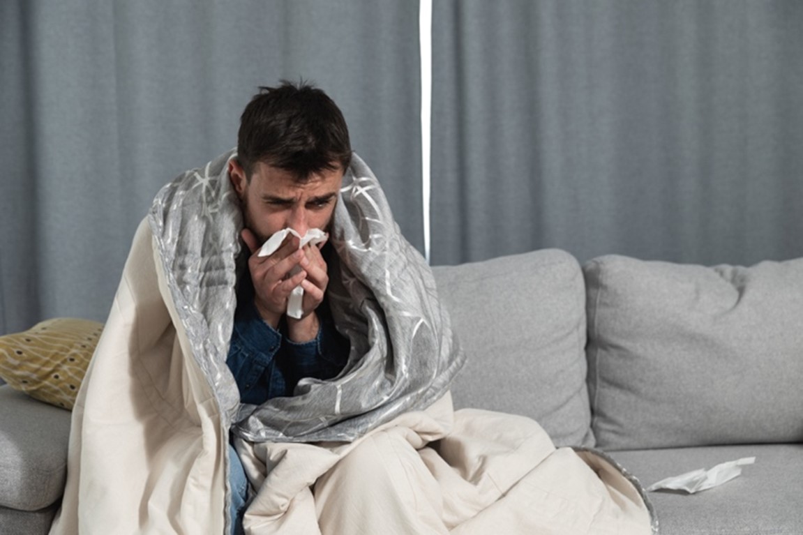 A man is sitting on a sofa wrapped in a blanket blowing his nose.