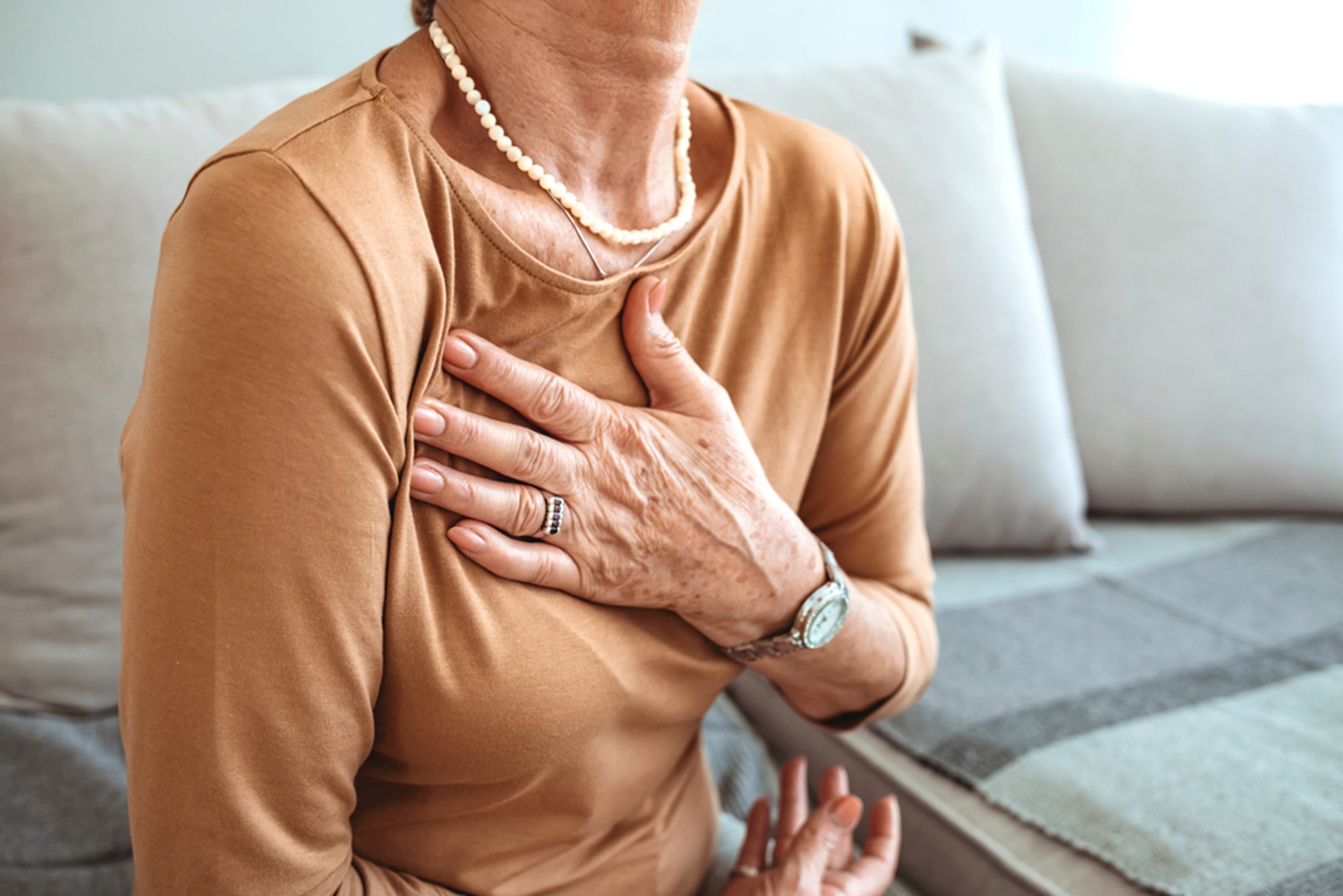 A closeup image of an older woman with her hand over her heart as she sits on the sofa.