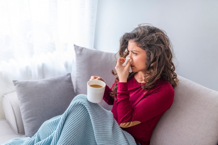 A woman in a long-sleeve maroon shirt sits on the sofa covered in a blanket, blowing her nose with one hand and holding a cup of tea with the other.