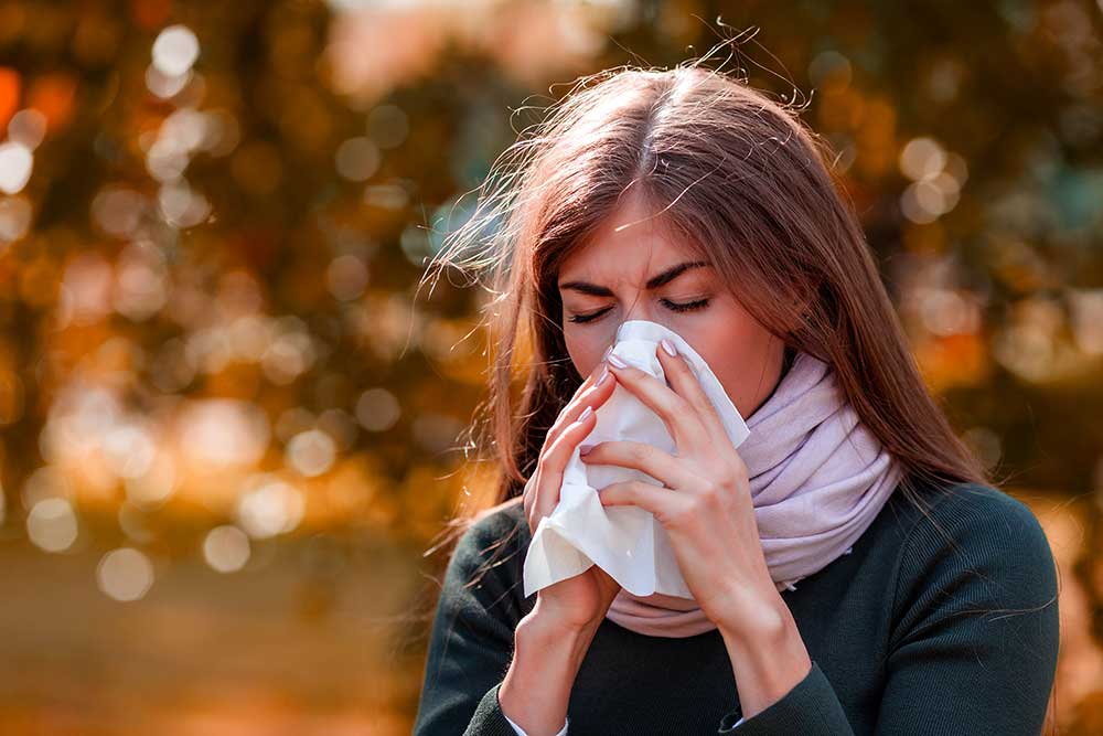 A woman outside on a fall day blows her nose with a tissue.