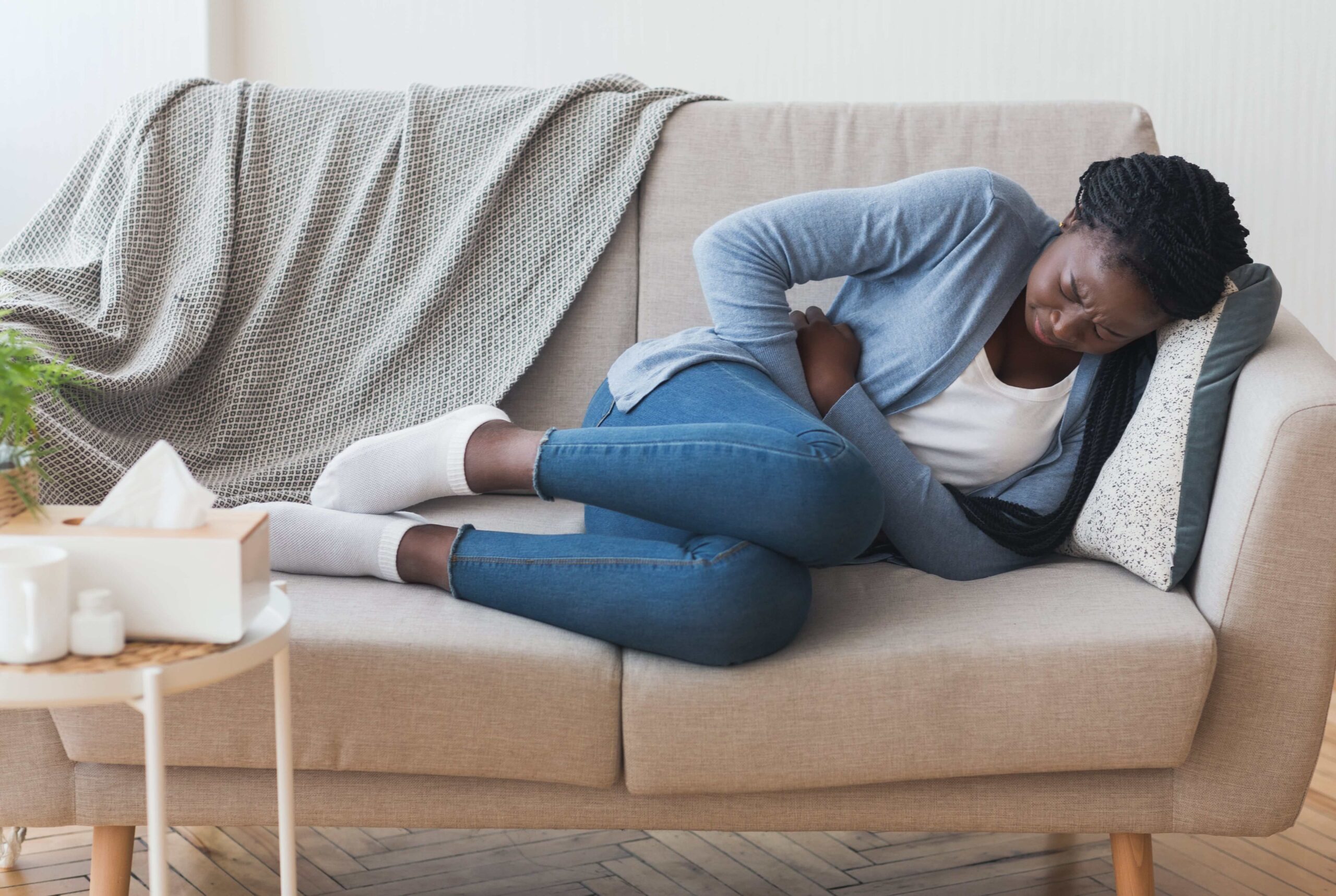 A woman is curled up on the sofa, holding her hands to her stomach and wincing in pain.