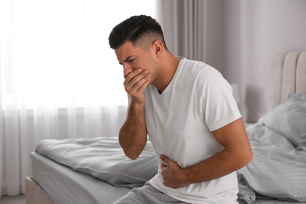 A man in a t-shirt sits on a bed, clutching his stomach and mouth. He appears nauseated.
