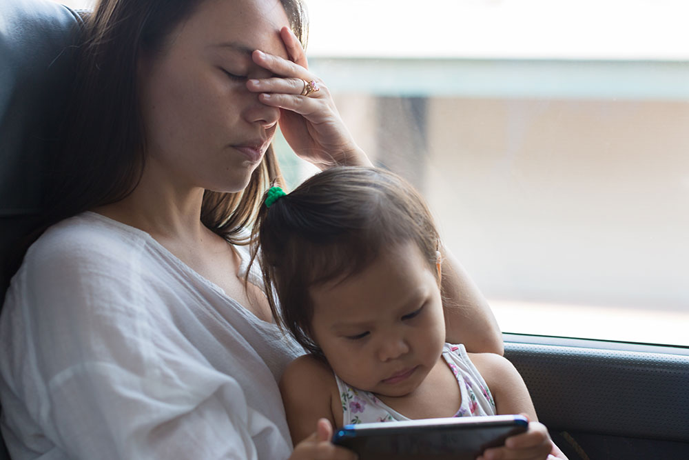 A woman with a young girl in her lap sits on a bus. The woman’s eyes are closed. She holds her forehead and appears sick.