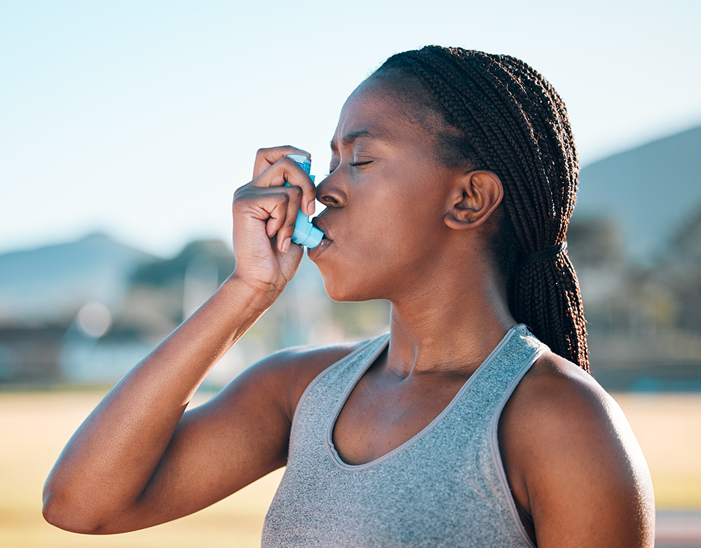 An African-American woman uses an asthma inhaler outdoors with her eyes closed.