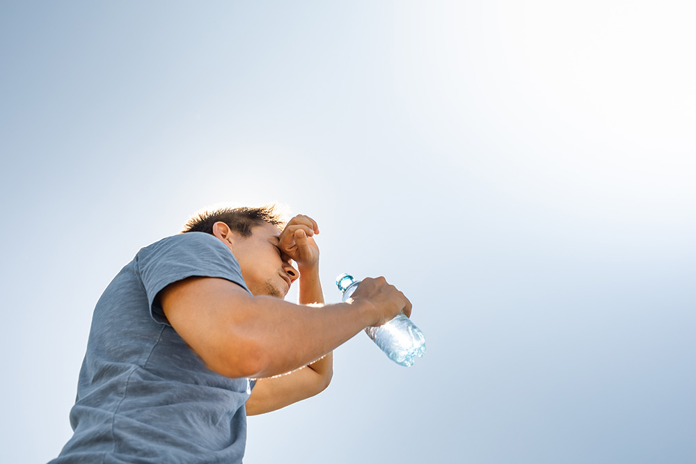 A man wearing a T-shirt is outside on a sunny day. He holds a water bottle in one hand and holds his other hand to his forehead.