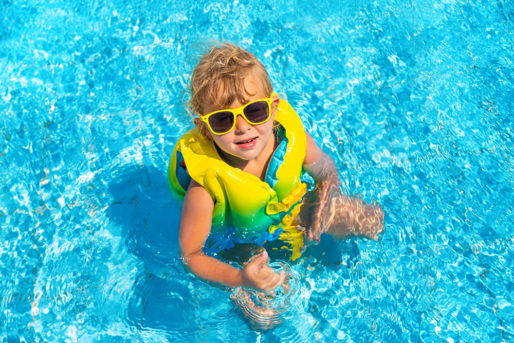 Small child wearing flotation device and sunglasses sitting in a shallow pool.