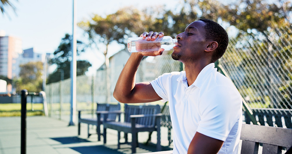 Black man in athletic wear drinking water from a bottle on an outdoor tennis court during a training break
